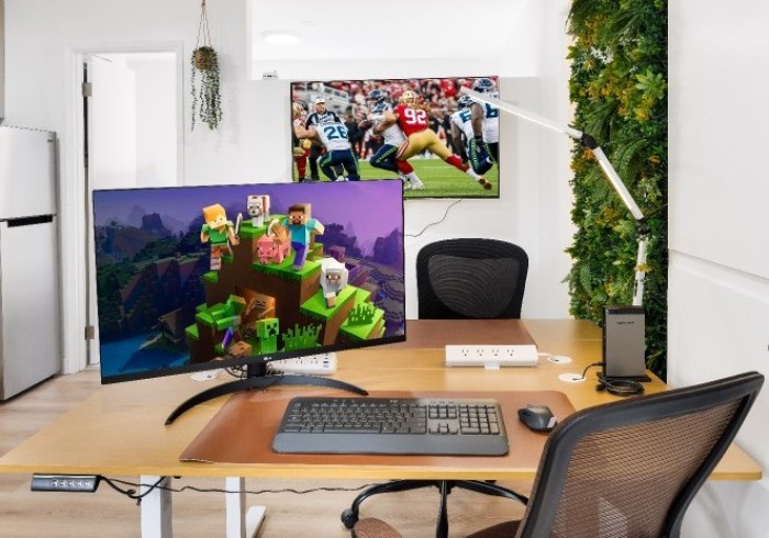 A large desk with monitor, keyboard, ergonomic chair, task lamp, and TV in the background.